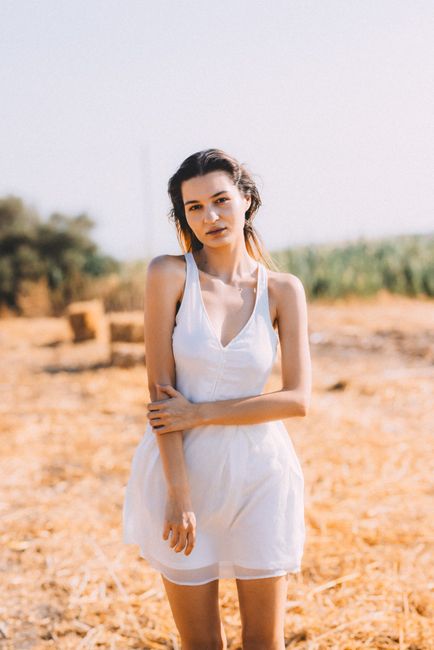 A young woman in a white dress stands in a sunlit field with hay bales.