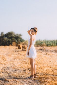 A young woman in a white dress stands in a sunlit field with a backdrop of trees.