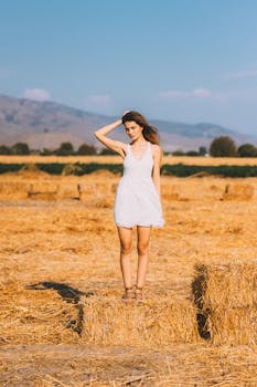 A young woman in a white dress stands on a straw bale in a sunlit hayfield with distant hills.