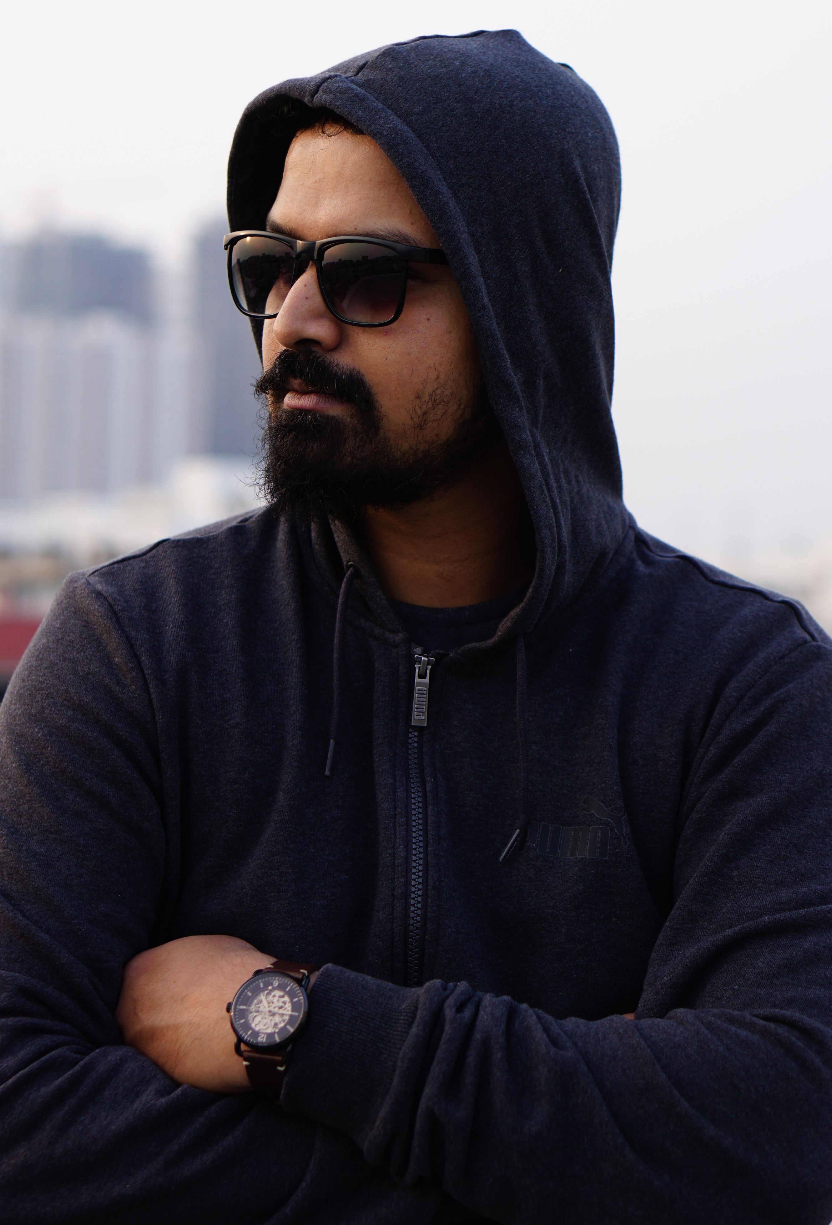 Bearded man with sunglasses and hoodie, exuding mystery in city backdrop.