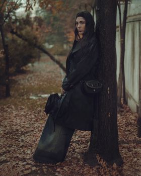 Moody portrait of a woman in a coat leaning against a tree in autumn.