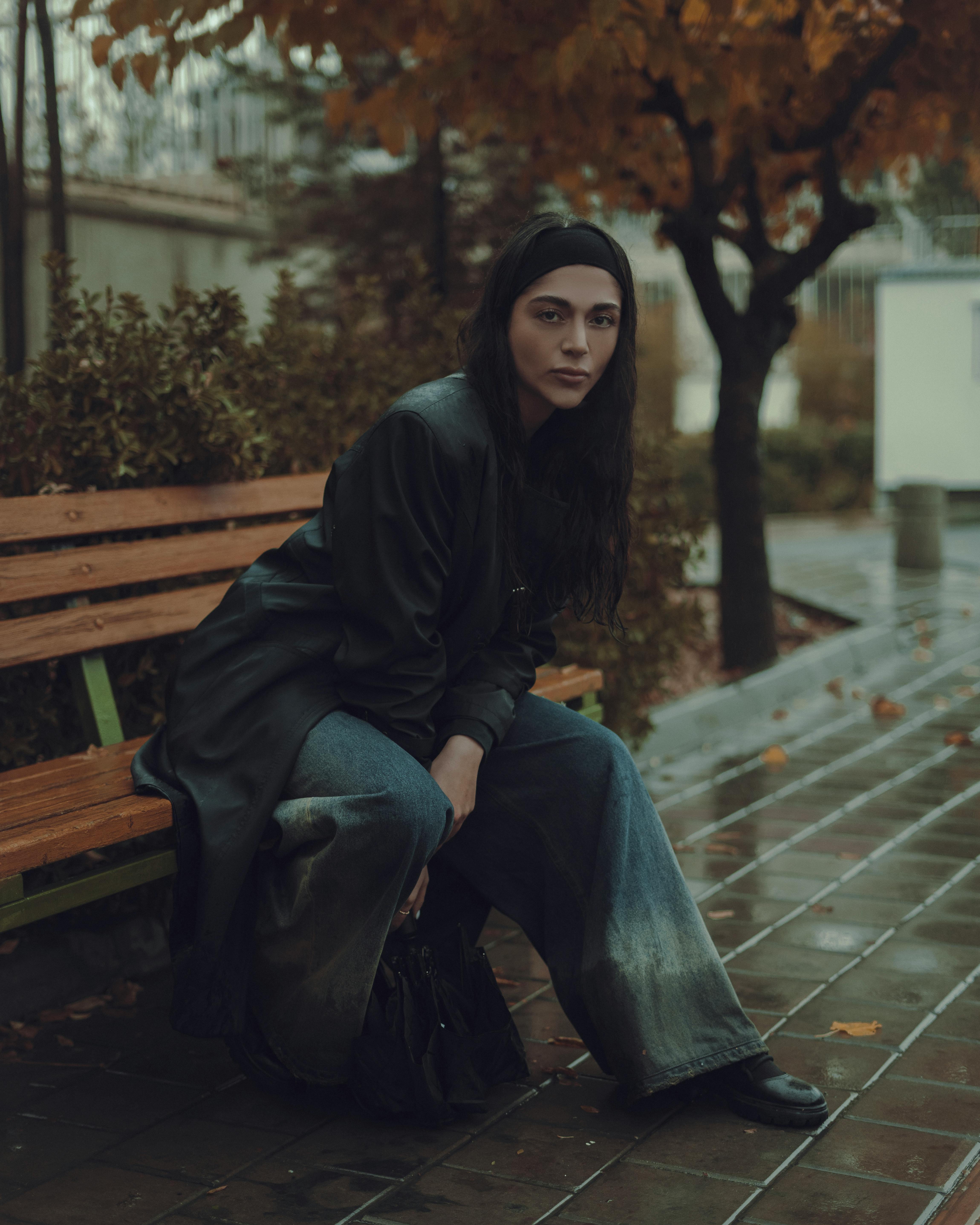 A woman sits alone on a park bench, surrounded by autumn leaves on a rainy day.