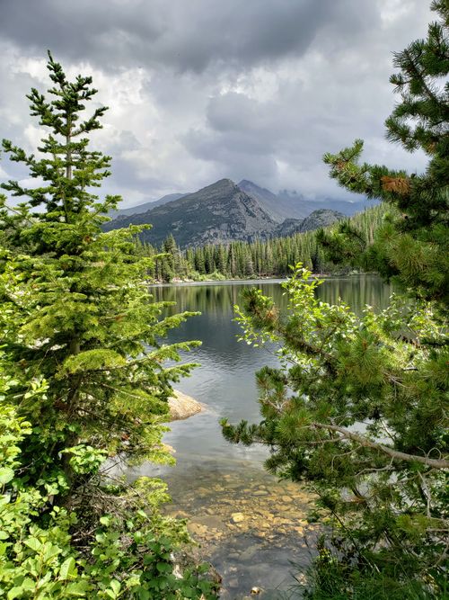 Free A serene mountain lake surrounded by lush greenery and pine trees under a cloudy sky. Stock Photo