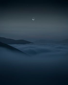 Ethereal night scene with crescent moon and stars above foggy mountain landscape.