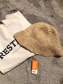 Straw hat, canvas bag, and sunscreen on sandy beach. Perfect summer essentials.