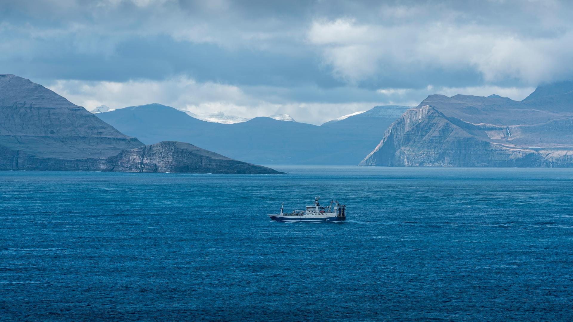 A scenic view of a fishing vessel navigating the deep blue sea near the Faroe Islands under a cloudy sky.