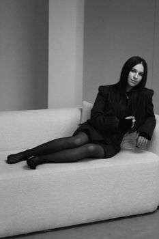 Woman in a black suit sitting on a white sofa, posing elegantly.