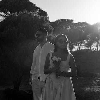 Stylish couple in monochrome setting among sunlit trees, exuding elegance.