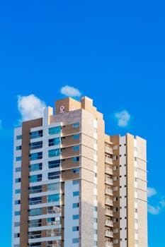 Modern high-rise building with glass balconies set against a bright blue sky with clouds.