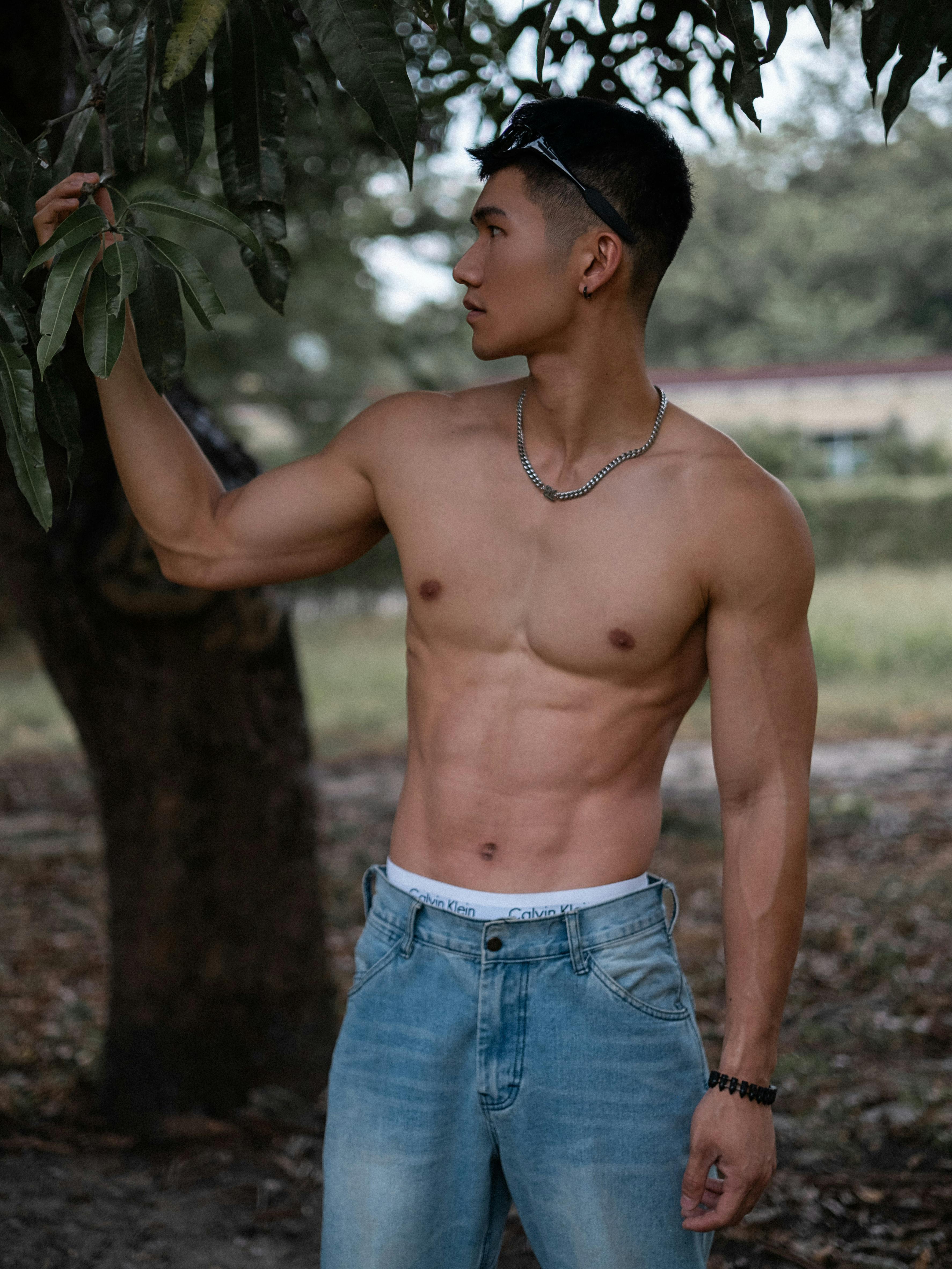 Shirtless young man in jeans posing outdoors, highlighting fitness and nature.
