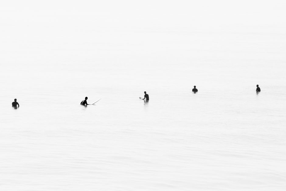 Silhouetted surfers waiting in calm ocean waters, creating a minimalist black and white scene.