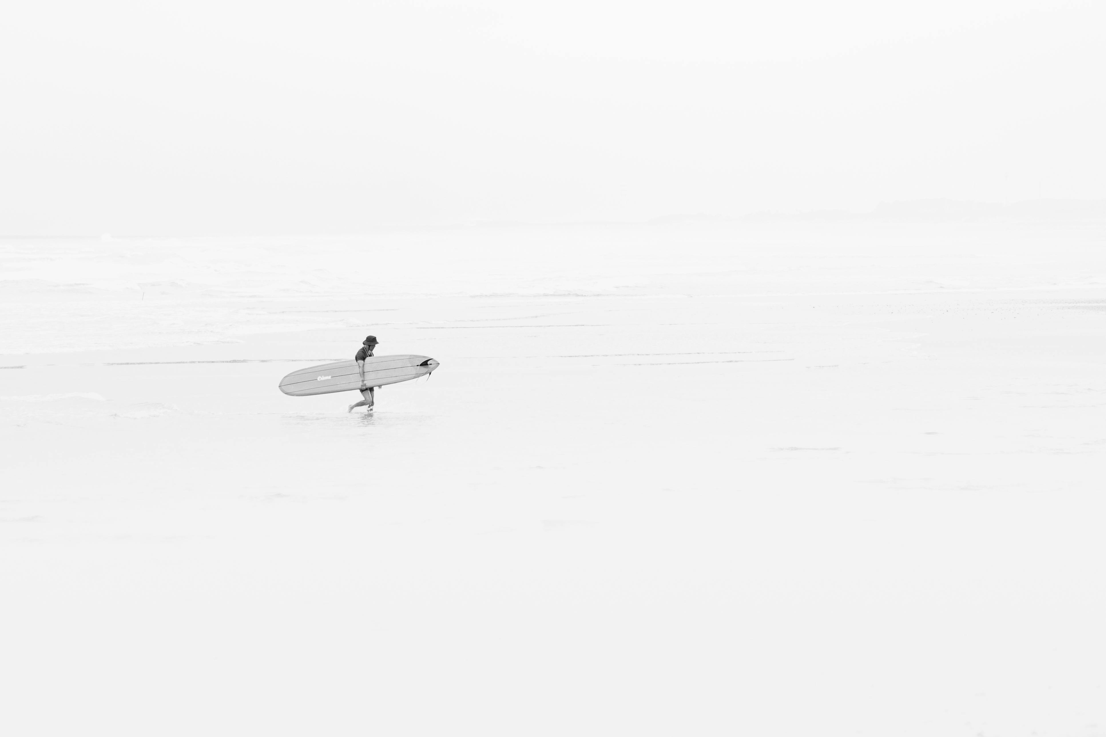 A solitary surfer walks along the serene Takanabe Beach, Japan, carrying a surfboard.