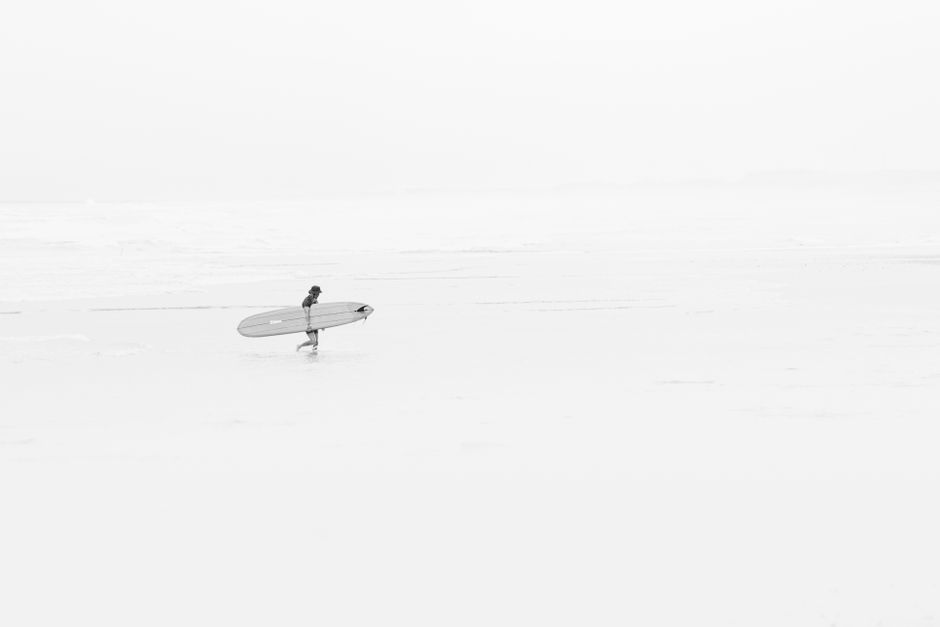 A solitary surfer walks along the serene Takanabe Beach, Japan, carrying a surfboard.