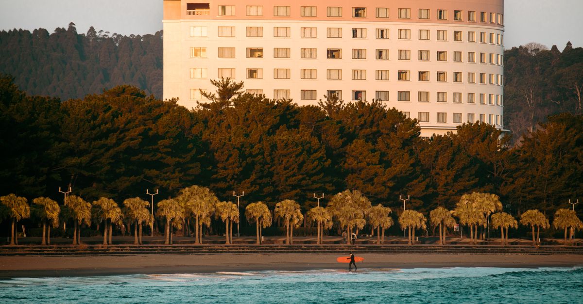 Surfer carrying board on Miyazaki Beach with hotel and sunrise backdrop in Japan.