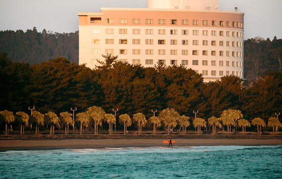 Surfer carrying board on Miyazaki Beach with hotel and sunrise backdrop in Japan.