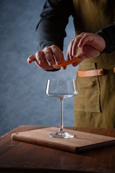 Close-up of bartender adding orange twist to a cocktail glass on a wooden board.