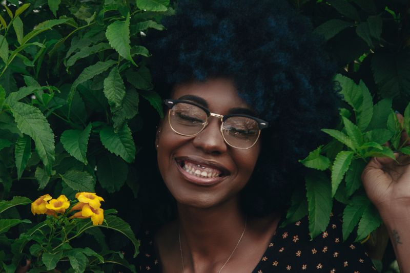 A Black woman smiles outdoors in daylight surrounded by greenery. More melanin means the skin makes less vitamin D from sun, so deficiency is common in Black adults.