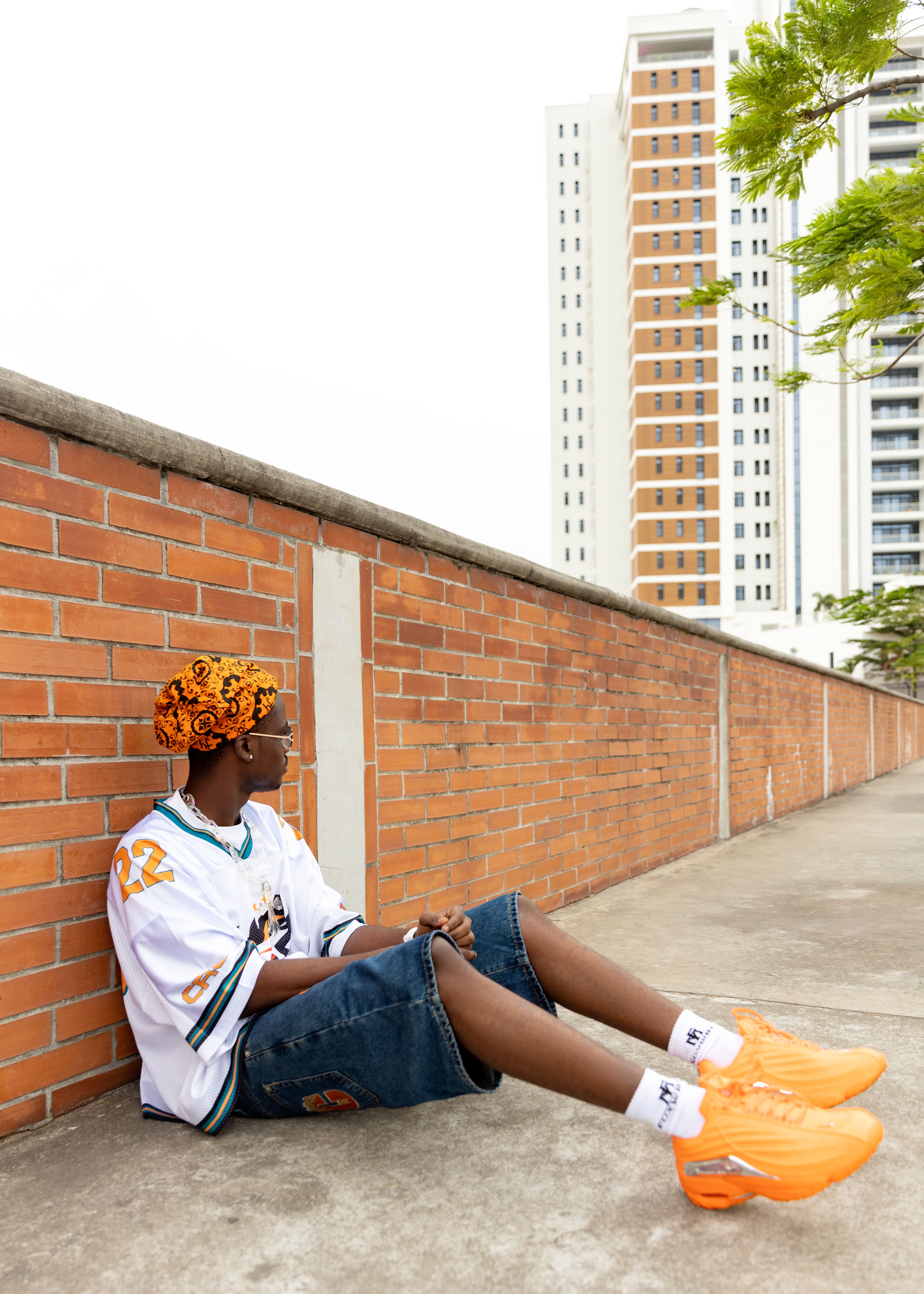 Young man in trendy outfit sitting against brick wall with high-rise buildings.