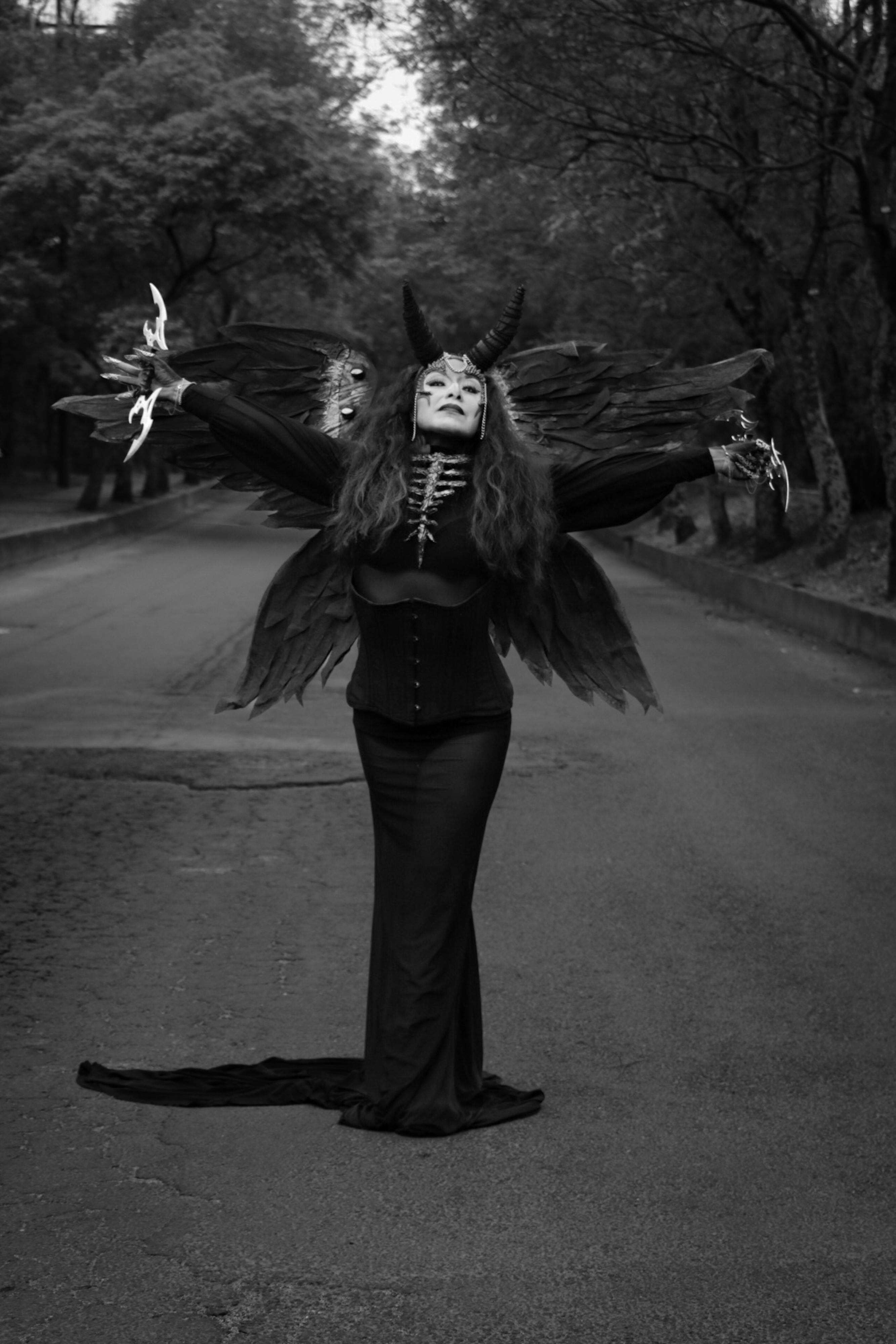 A person in a dramatic gothic costume stands in a forested road.