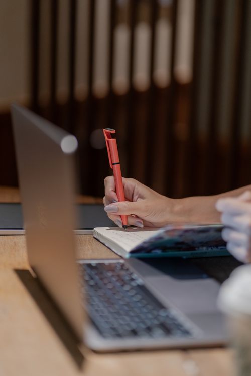 Person Writing in Notebook at Laptop Desk · Free Stock Photo