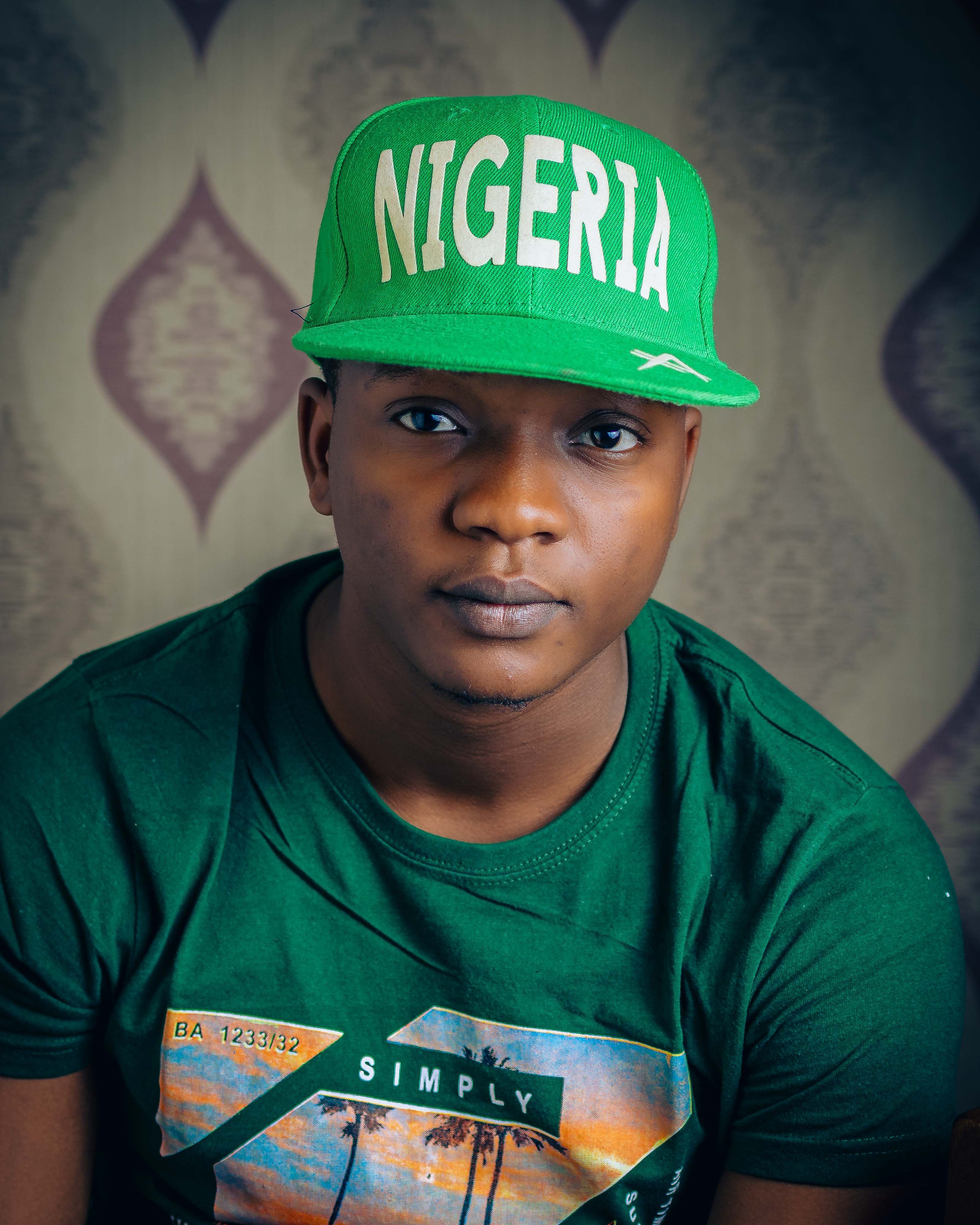 A young man wearing a green Nigeria cap and T-shirt against a patterned background.