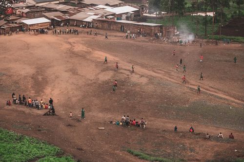 Free Children play soccer on a rural field while villagers watch. Aerial shot capturing community life. Stock Photo