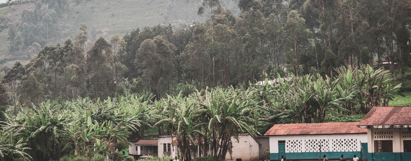 Children play soccer on a grassy field surrounded by lush African landscape and small village buildings.