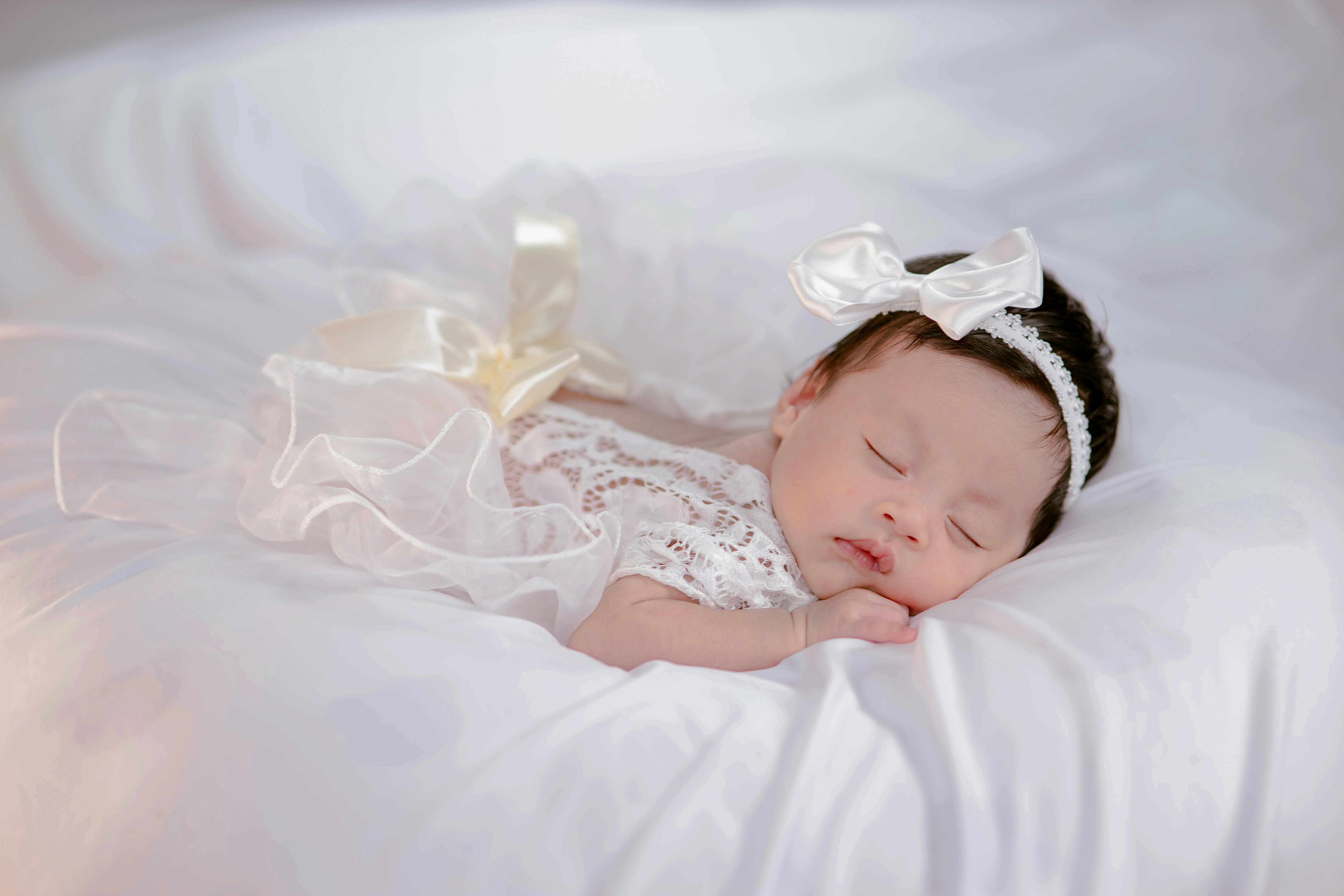Free Peaceful sleeping newborn wearing white lace and bow, lying on soft bedding. Capturing the serenity and innocence of infancy. Stock Photo