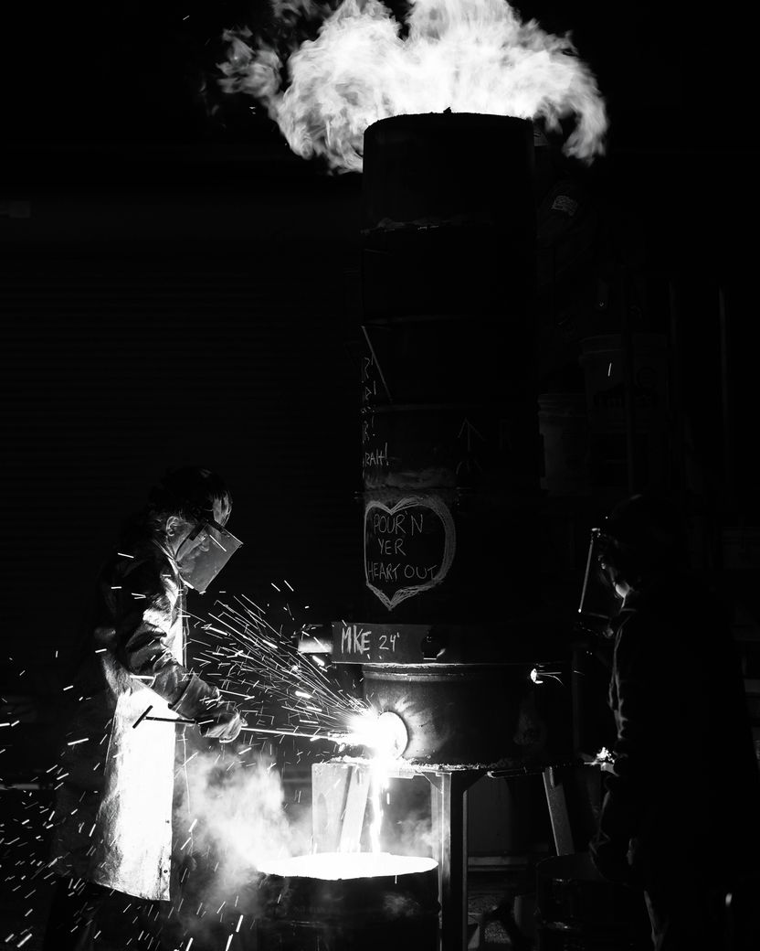 A black-and-white foundry scene at night, with flame breaking from the top of a tall cylindrical furnace and a worker in protective gear tapping molten metal into a crucible amid sparks.