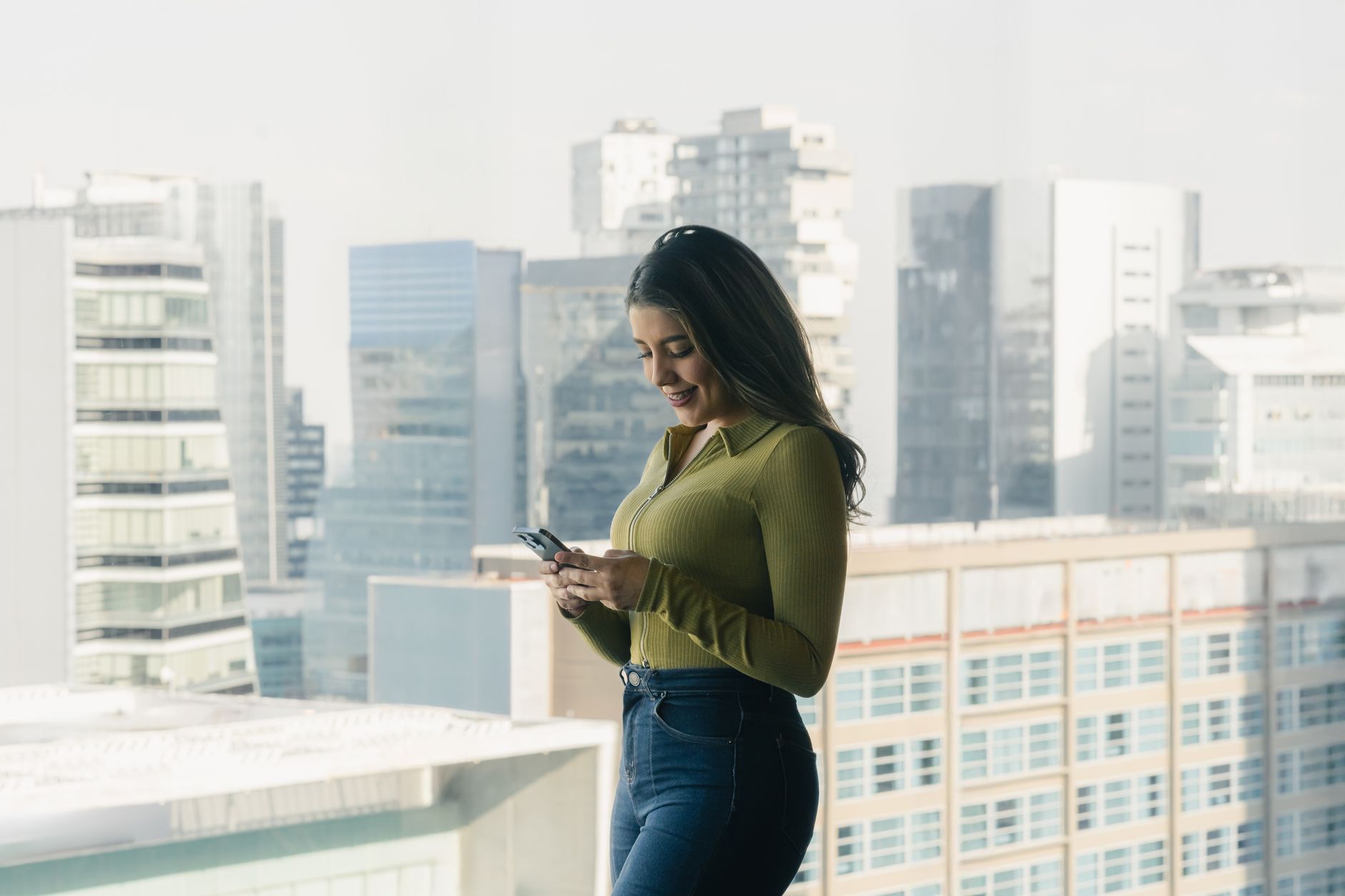 Woman looks at smartphone inside high-rise building with cityscape in background.