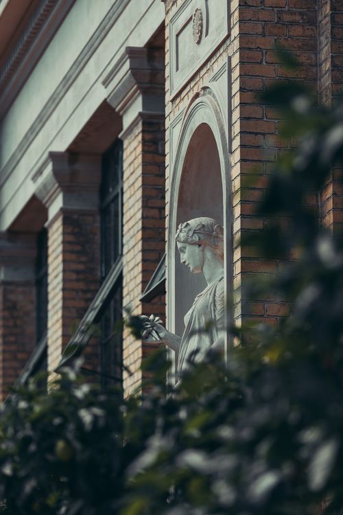 Free A beautiful classical statue adorns a brick building facade with lush greenery. Stock Photo