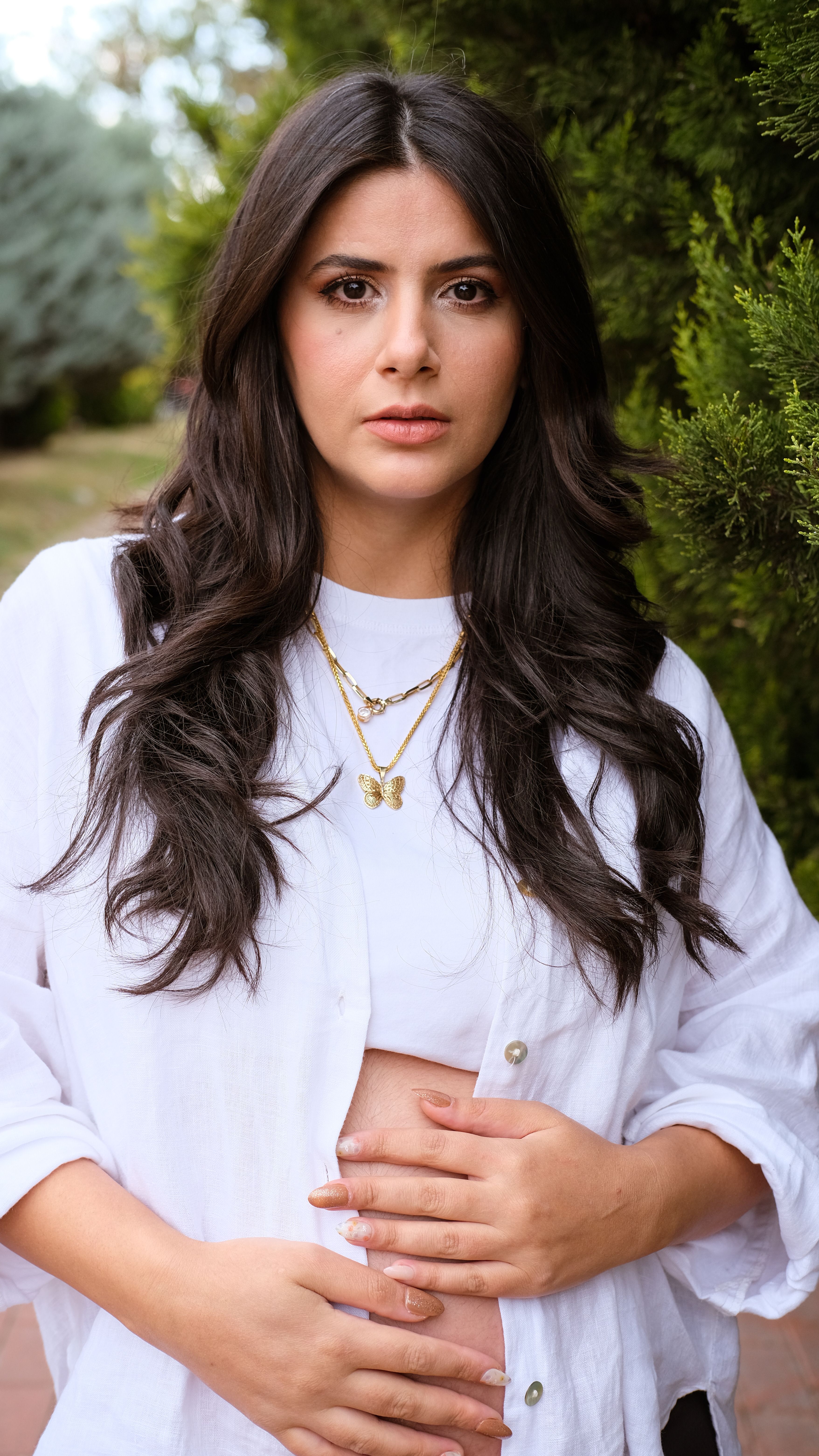 Portrait of a young woman with long hair in a white outfit posing outdoors against greenery.