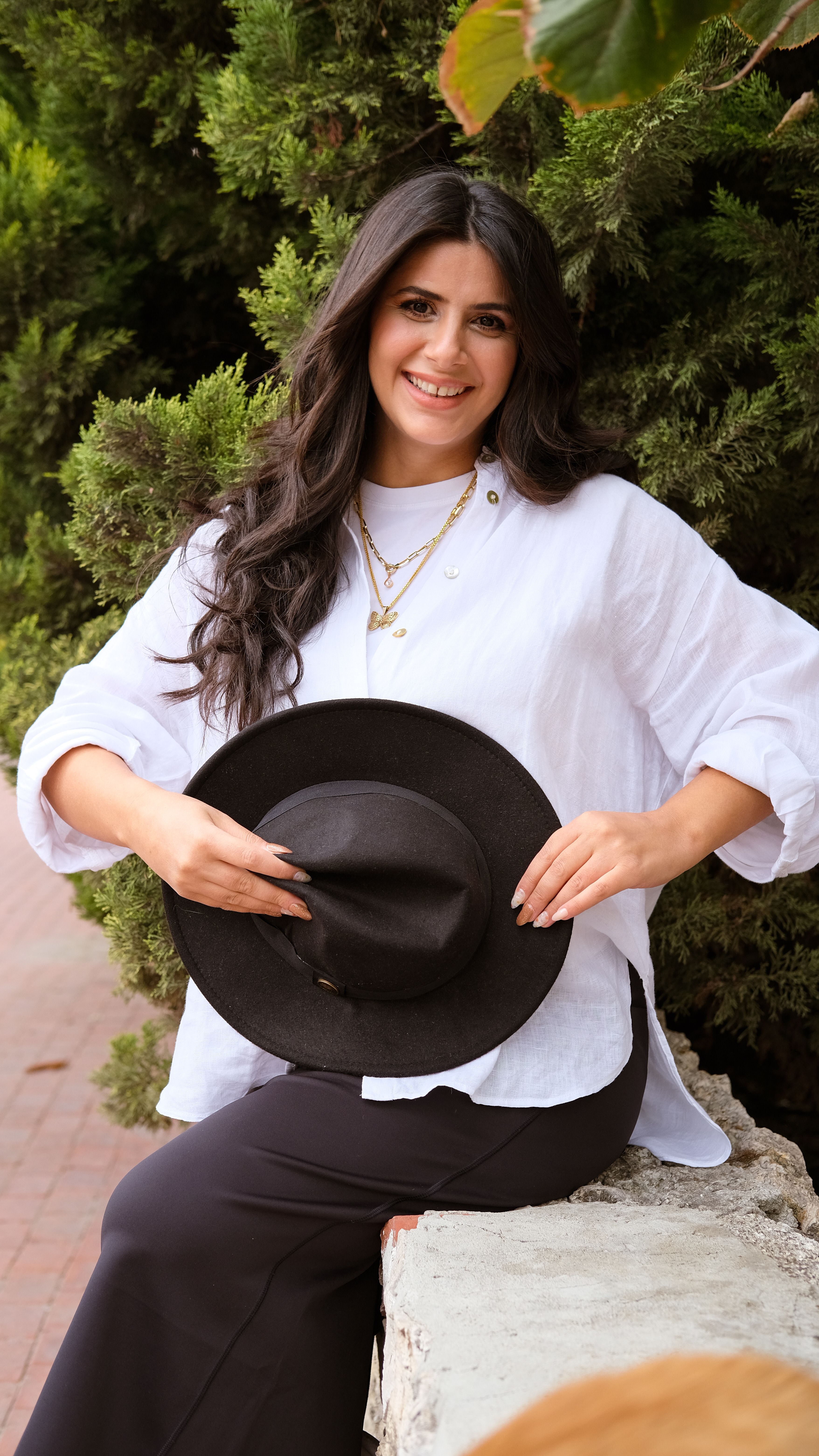 Smiling young woman in casual wear holding a hat, outdoors against green foliage.