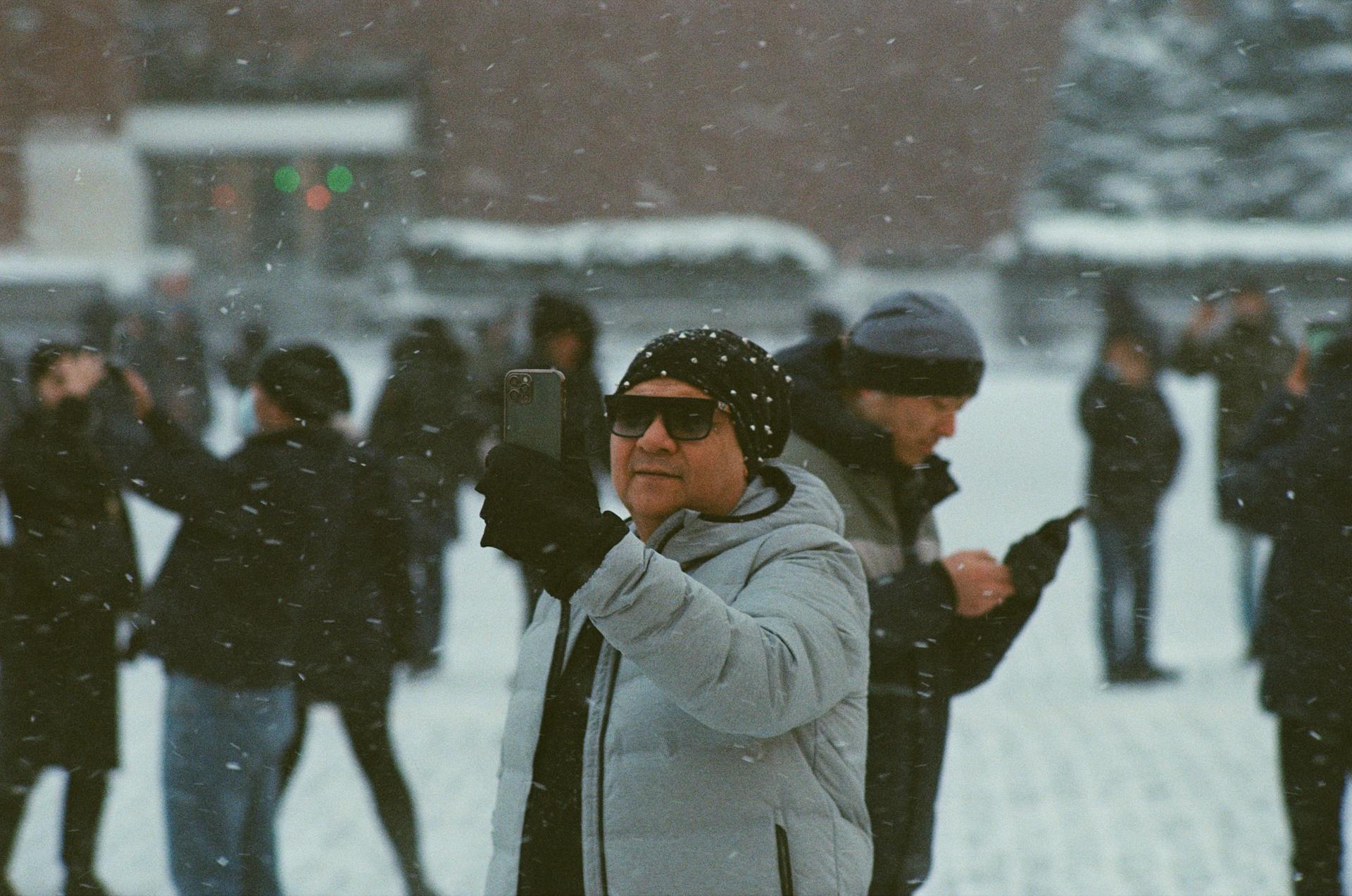 A person takes a selfie in a snowy city scene, capturing a winter moment with onlookers.