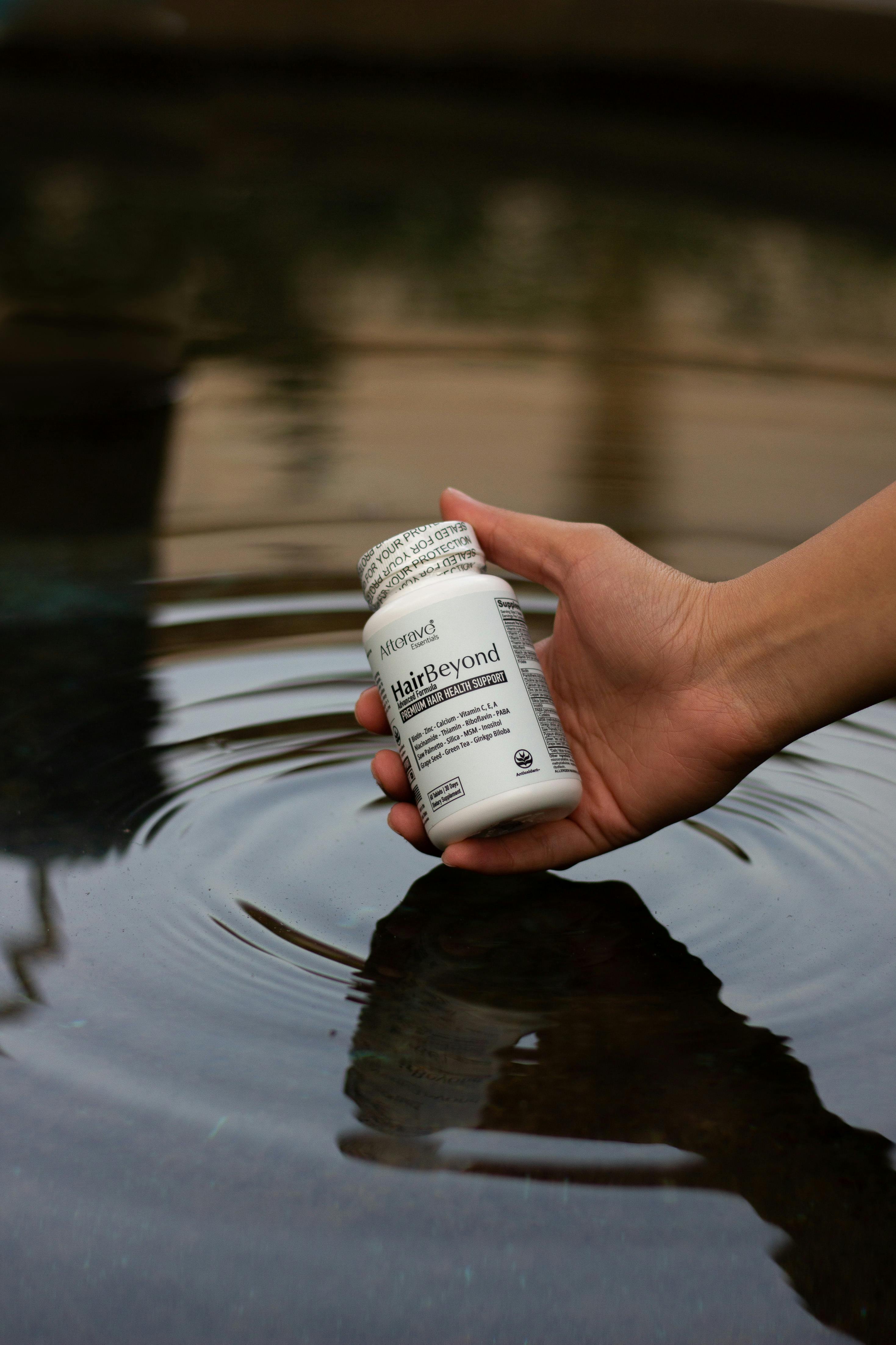 A hand holding a bottle of HairBeyond supplement above reflective water, creating ripples.