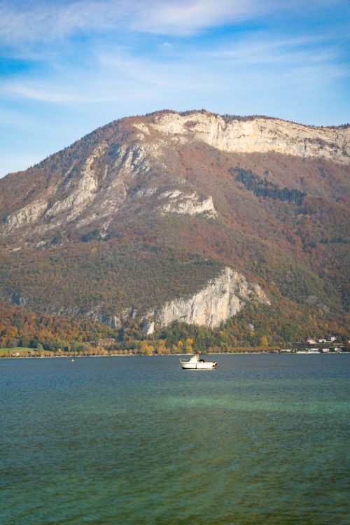 Free A serene view of Lake Annecy with a lone boat against a backdrop of autumnal mountains. Stock Photo