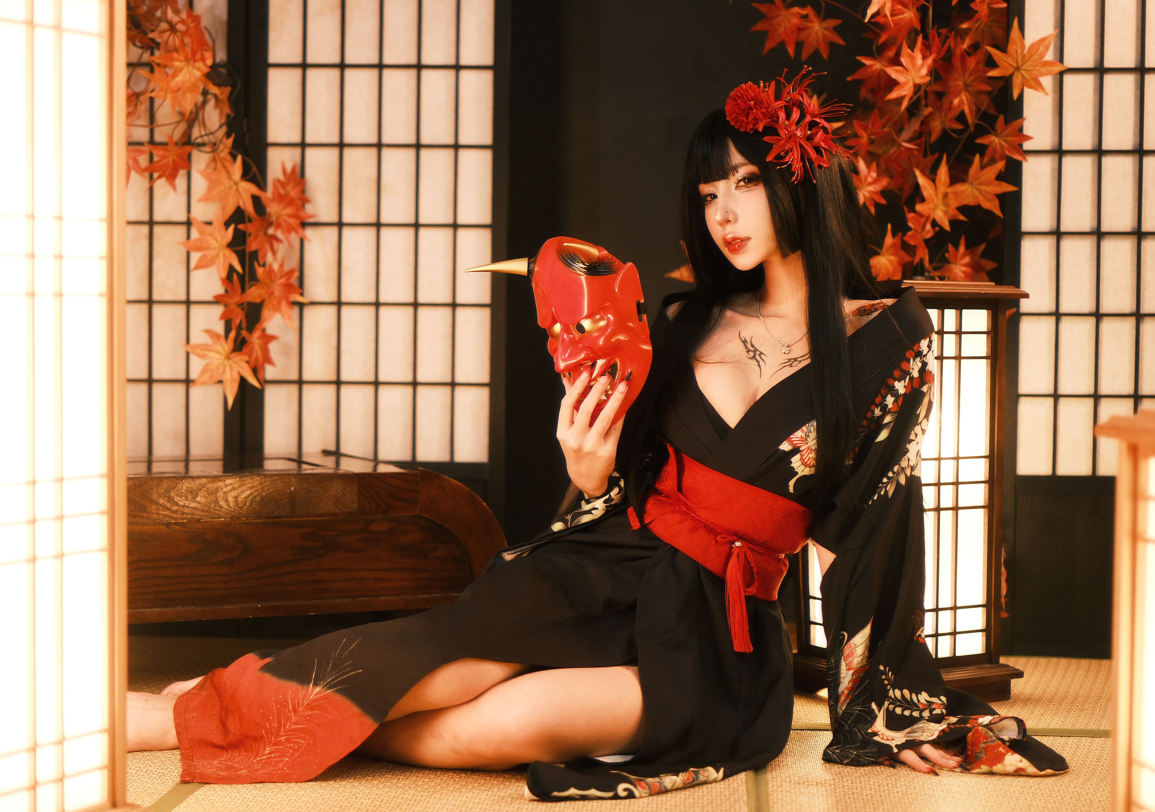A woman in a traditional Japanese kimono holding a red mask indoors with autumn decor.