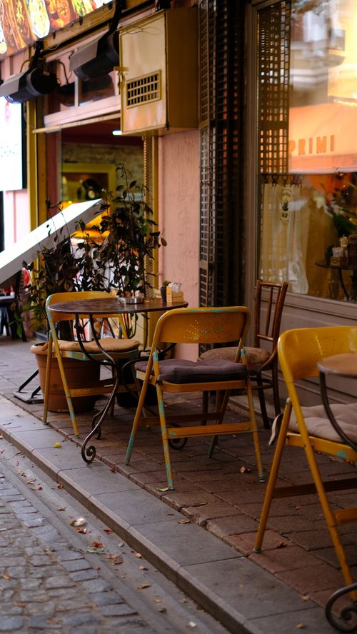 Free Cozy outdoor cafe scene with yellow chairs and tables on a bustling city street. Stock Photo
