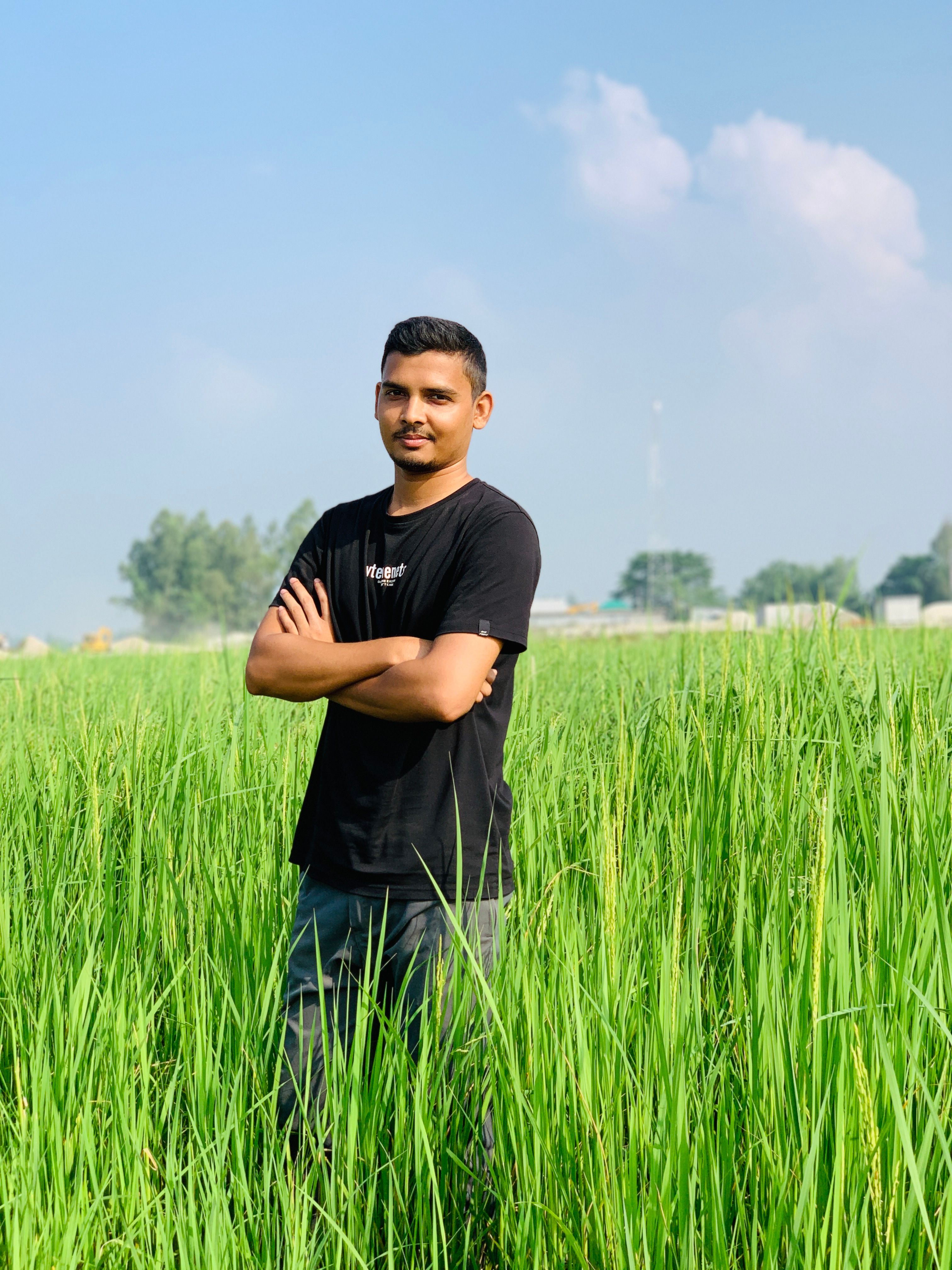 A young South Asian man stands confidently amidst lush green rice paddies under a clear blue sky.