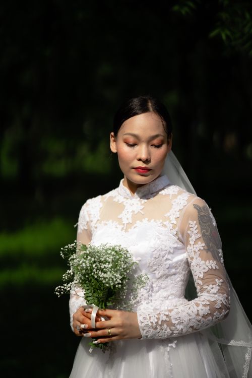 Free Graceful bride in white lace dress holding a bouquet and standing outdoors. Stock Photo