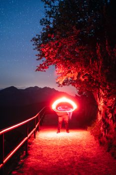 A person creates vibrant red light trails on a night hike in Bavaria, Germany, under a starry sky with mountains in the background.