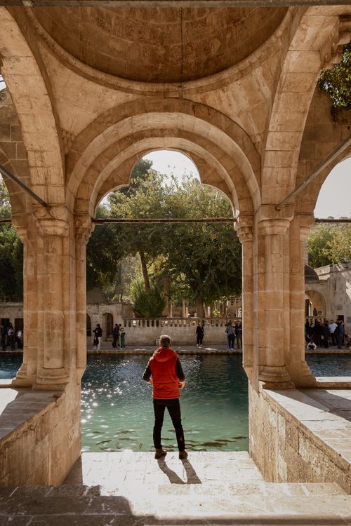 Free A person in a red jacket looks out over a serene pool framed by a historic stone archway. Stock Photo