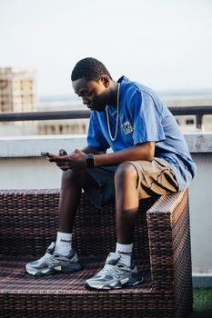 A young man in casual attire texts on a rooftop terrace, enjoying a sunny day.