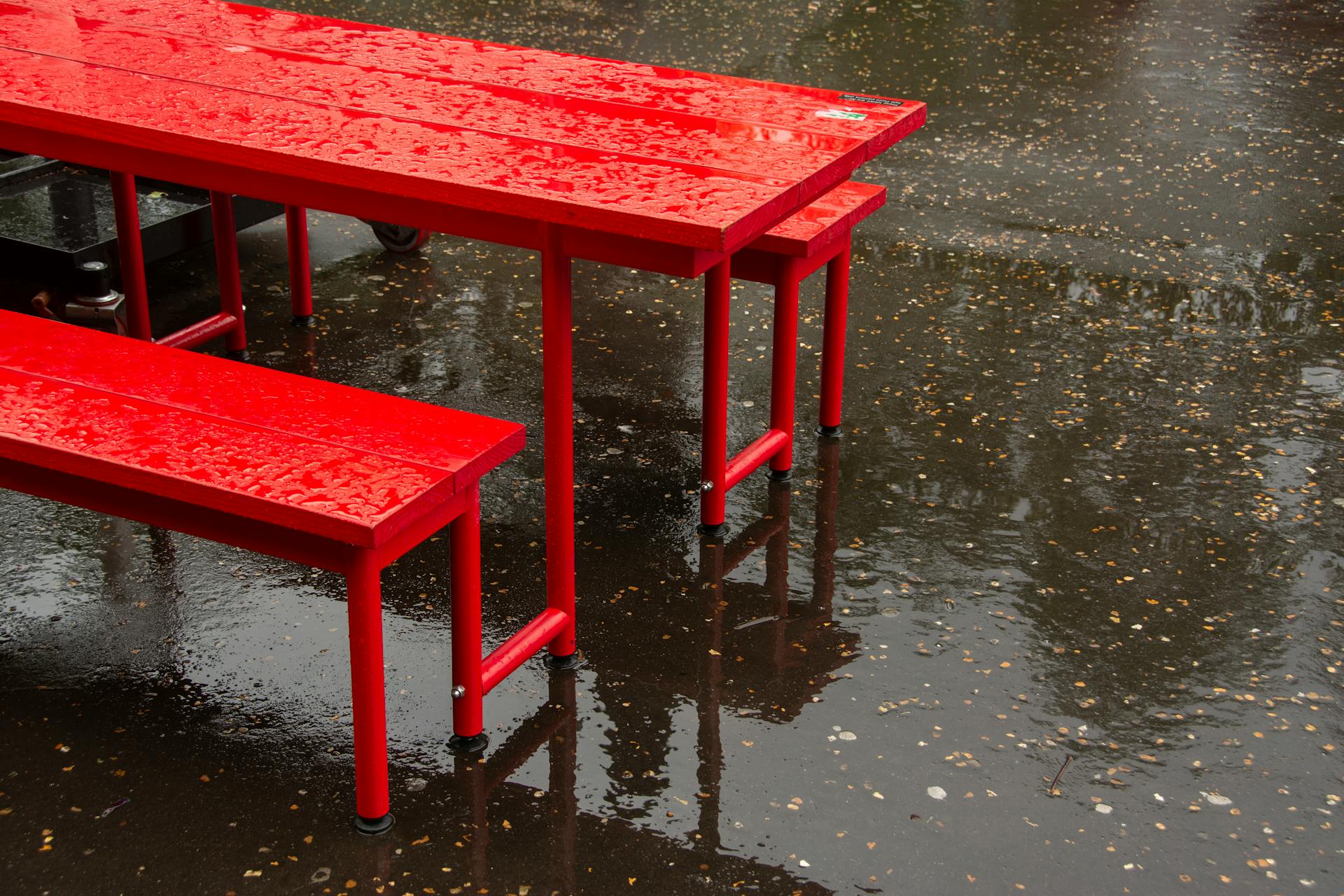 Bright red table and bench set on wet asphalt with rain droplets.