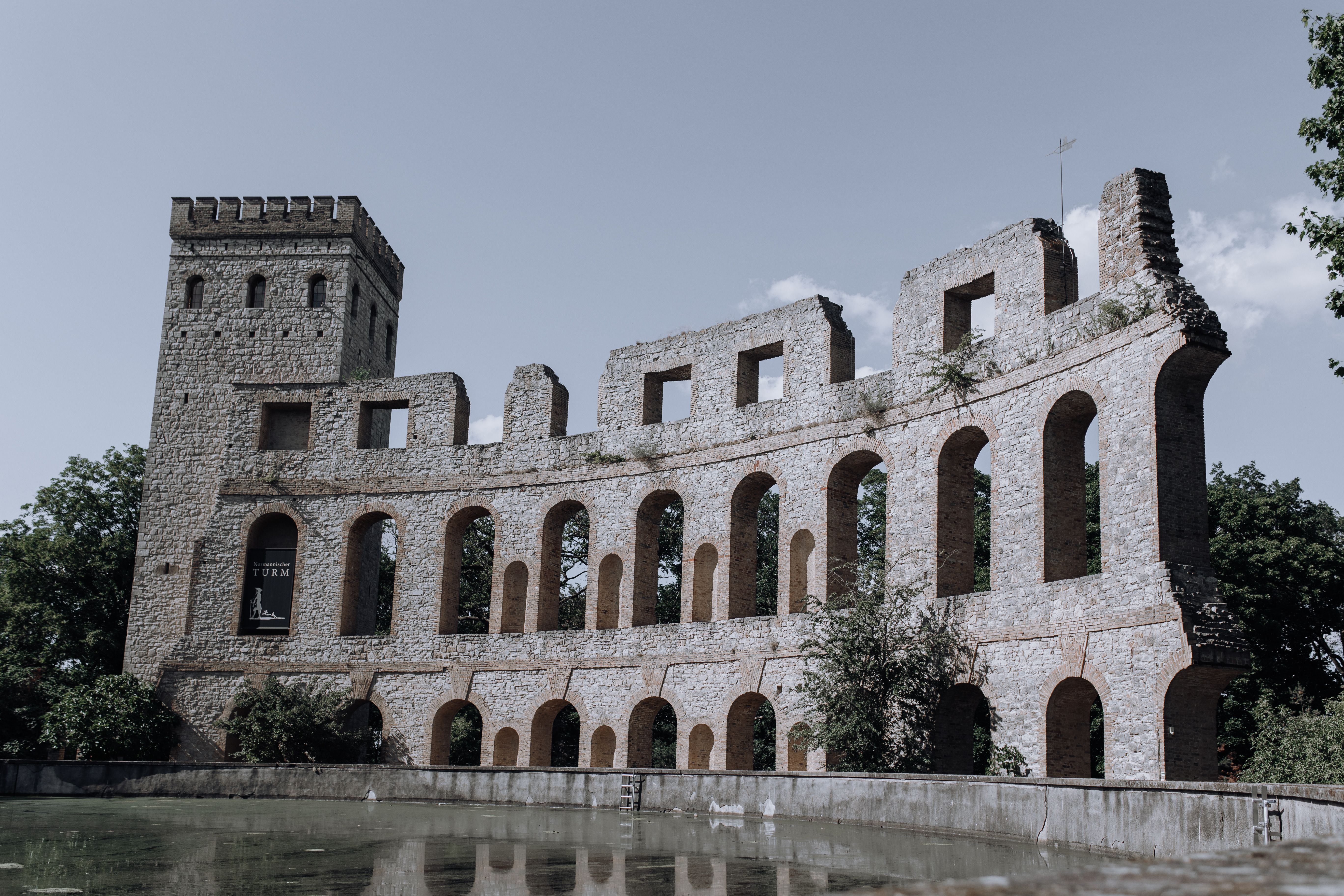 Norman Tower and stone arcade ruins in Potsdam, Germany, beside a reflecting pool in bright daylight with no people visible