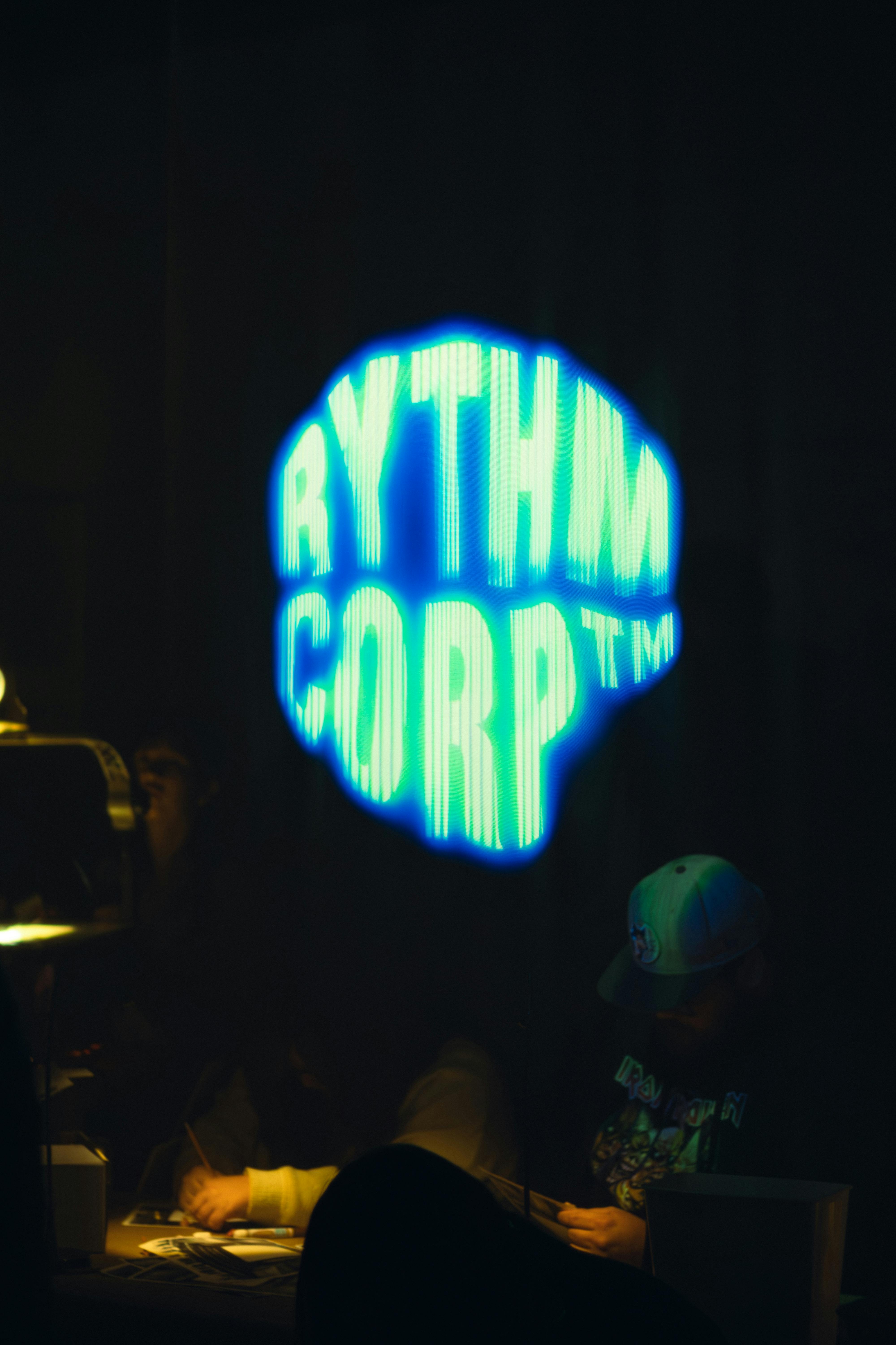Kostenlos Neonblaue Typografie „RYTHM CORP“ in schwach beleuchteter Umgebung mit sichtbaren Personen bei Aktivitäten. Stock-Foto