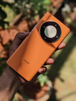 A close-up of a hand holding an orange HONOR smartphone with a circular camera module.