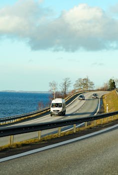 A picturesque coastal road with vehicles, overlooking the ocean and under a partly cloudy sky.