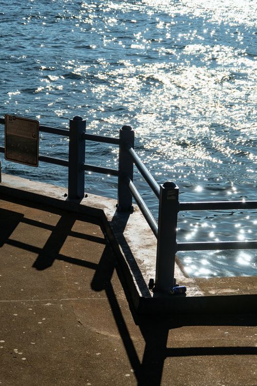 Free A sunlit railing casts shadows over a sparkling water surface on a sunny day. Stock Photo