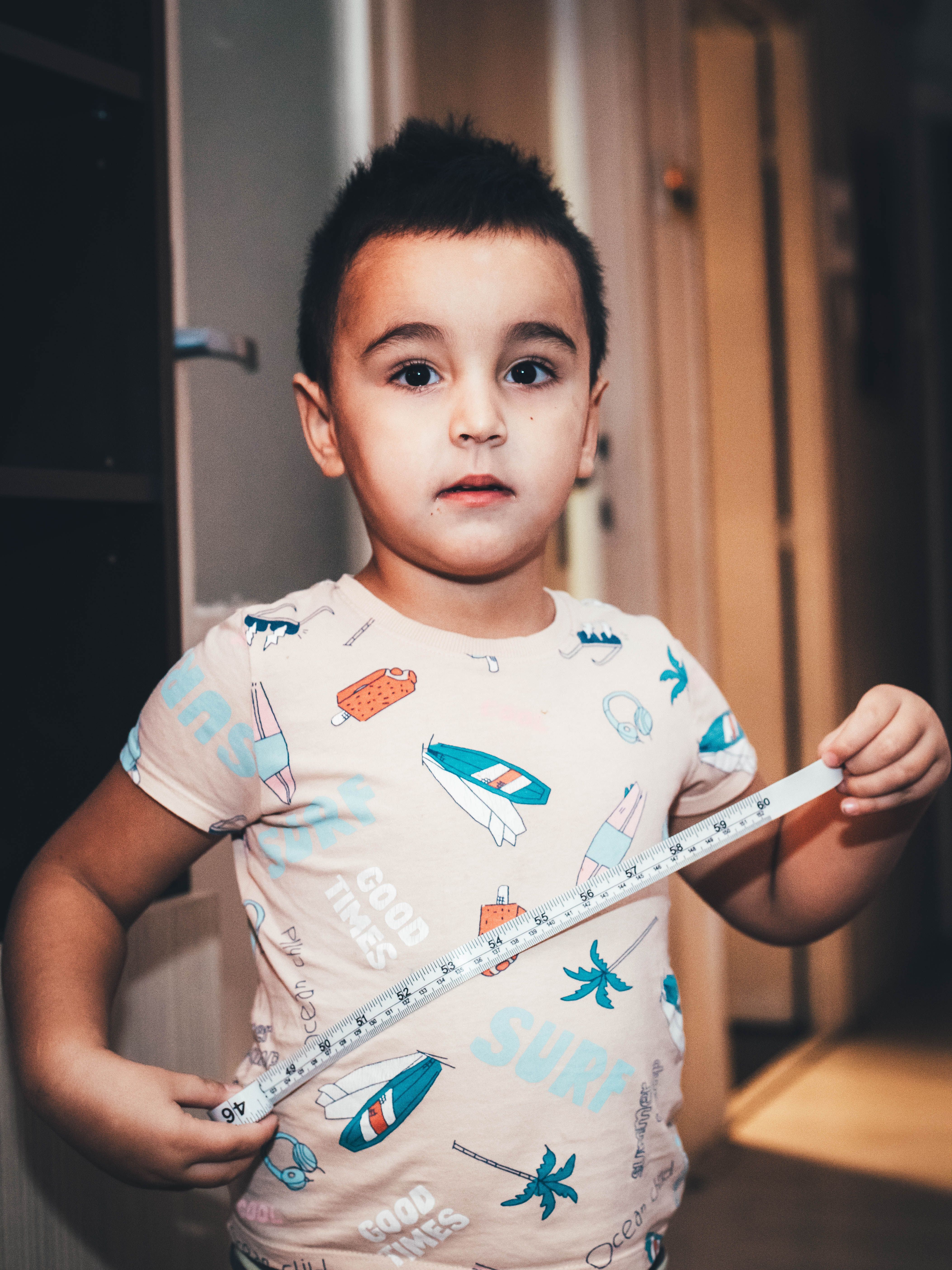 A young child holds a measuring tape indoors wearing a playful t-shirt with surf designs.